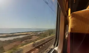 Blick aus dem Zugfenster auf eine Küstenlandschaft mit Meer und Bahngleisen. Die Reise mit dem Zug nach Mallorca beginnt.
