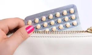 Dłoń z różowym lakierem wyciąga blister z tabletkami antykoncepcyjnymi z białej kosmetyczki.