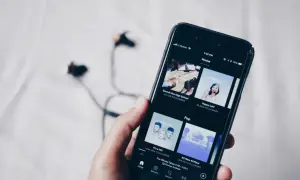 Dłoń trzyma smartfon z aplikacją do streaming muzyki. W tle leżą słuchawki.