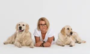 Kobieta w okularach leży między dwoma golden retrieverami na białym tle.
