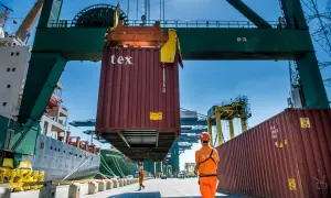 Ein Container wird im Antwerpener Hafen von einem Kran gehoben. Arbeiter in orangefarbener Kleidung überwachen die Aktion.