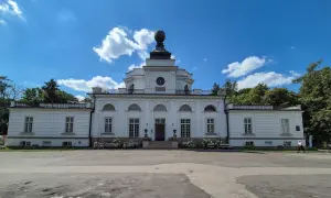 Pałac w Jabłonnie, biała, klasycystyczna budowla z charakterystyczną kopułą, otoczona zielenią, pod błękitnym niebem z chmurami.
