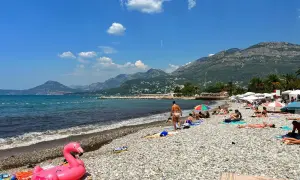 Słoneczny dzień na plażach Zatoki Kotorskiej. Ludzie odpoczywają na kamienistym brzegu, a w wodzie widać dmuchanego flaminga.