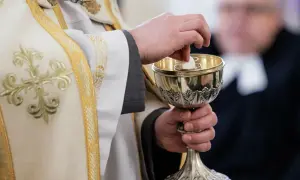 Dłoń księdza w białej szacie z haftem podaje hostię do kielicha. Komunia bez spowiedzi, symbol wiary.