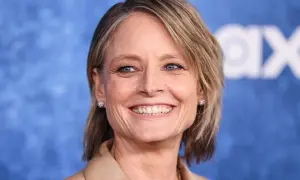 Uśmiechnięta Jodie Foster, gwiazda wielu niezapomnianych **jodie foster filmy**, w beżowym żakiecie i z błyszczącymi kolczykami.