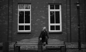 Młoda kobieta siedzi na ławce przed ceglanym budynkiem z dużymi oknami. To klasyczny plan amerykański w fotografii, podkreślający jej postać w otoczeniu architektury.