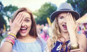 Dwie dziewczyny z brokatem na twarzy, zakrywające jedno oko dłonią, cieszą się atmosferą festiwalową.