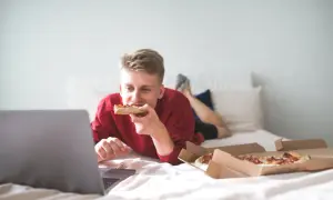 A young man eats pizza in bed while watching a laptop. Is it bad to go to sleep after eating? This scene suggests a late-night snack before bed.