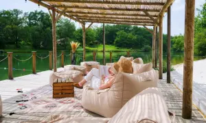 Détente sous le toit de chaume du whaka lodge, au bord d'un lac paisible. Une femme lit, entourée de poufs confortables.