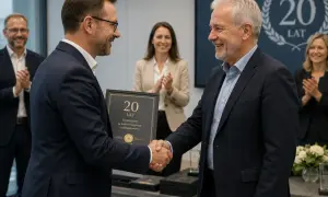 Wręczenie nagrody jubileuszowej za 20 lat pracy. Dwóch mężczyzn w garniturach ściska sobie dłonie, jeden z nich trzyma dyplom.