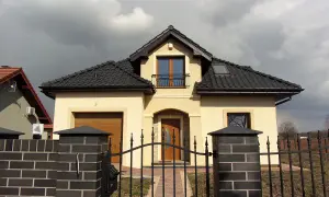 Jasny dom z brązowym dachem, bramą garażową i wejściem, ogrodzony czarnym płotem.