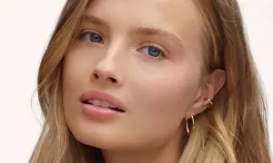 Visage d'une jeune femme aux yeux bleus et aux lèvres rosées, avec un maquillage subtil, style "makeup no makeup". Elle porte des boucles d'oreilles dorées.