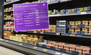 Fioletowa plansza z cenami masła w promocji na tle półek sklepowych z produktami.
