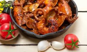 Un plat de calamars à l'armoricaine mijoté dans une sauce rouge, servi dans une poêle en fonte, avec des tomates et de l'ail à proximité.
