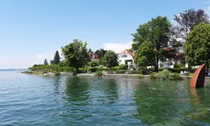 Die Hagnau Promenade am Bodensee mit weißen Häusern, grünen Bäumen und einer rostigen Skulptur im Wasser.