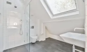 Moderno baño en buhardilla con ducha de cristal, inodoro y lavabo bajo una claraboya.