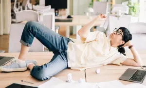 En person ligger utsträckt på ett skrivbord, omgiven av papper och laptops. Klädd i jeans och en ljus skjorta, ser hen ut att fundera över en papperslapp, en bild av en dysfunktionell arbetsplats.