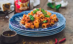 En tallrik med rigatoni all'arrabiata, toppad med basilika och parmesan. Bredvid syns en burk Mutti-tomatsås, vitlök och chilifrukter.