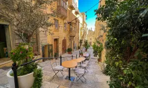 Malta: co warto wiedzieć? Urokliwa uliczka z kawiarnianymi stolikami i krzesłami, otoczona kamiennymi budynkami i zielenią.