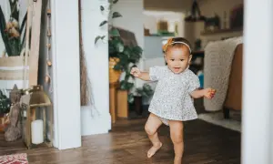 Radosne dziecko w kwiecistej sukience robi pierwsze kroki, kiedy dziecko zaczyna wstawać. W tle domowe wnętrze z roślinami.