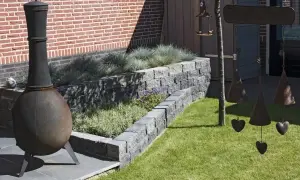 Ein Garten mit einem rustikalen Ofen und einer schönen Mauer aus **Pflanzsteinen setzen** für die Bepflanzung. Dekorative Glocken hängen von einem Baum.
