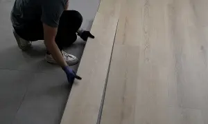 Persona con guantes instala piso SPC de madera clara sobre suelo gris.