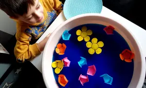 Chłopiec w żółtej bluzie obserwuje kolorowe papierowe kwiatki unoszące się na wodzie w misce.