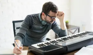 Mężczyzna zamyślony nad nutami, z ręką na czole, obok klawiatury. Chce zacząć grać na piano.