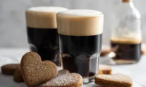 Två glas mörkt kaffe Karlsson med fluffigt skum, omgivna av hjärtformade pepparkakor pudrade med florsocker.
