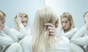 Blondynka w białej bluzie trzyma się za głowę, patrząc w lustro, gdzie odbija się jej zmartwiona twarz. Może to symbolizować walkę z chorobą psychiczną, gdzie kwetiapina jest jednym z leków.