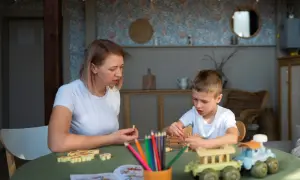Kobieta i chłopiec grają w drewniane gry na zielonym stole. W tle widać wystrój pokoju, przypominający salę w przedszkolu niepublicznym.