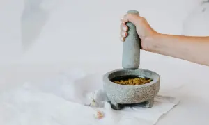Dłoń uciera składniki w moździerzu, pokazując, jak zrobić pesto. Obok leżą ząbki czosnku.