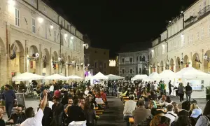 Serata di festa in una piazza delle Marche, con tavoli all'aperto e gente che mangia e socializza.