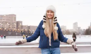 Uśmiechnięta blondynka w granatowej kurtce i białej, futrzanej czapce z pomponami. Idealny zestaw na lodowisko.