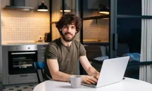 Uśmiechnięty mężczyzna z kręconymi włosami pracuje na laptopie w kuchni. Myśli o tym, jak zapłacić czynsz, ale jest zrelaksowany.