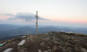 Szczyt góry z krzyżem i ławkami, w oddali widać pasma górskie. W tle zachodzące słońce i księżyc. Wokół rośnie trawa i pojedynczy smerek.