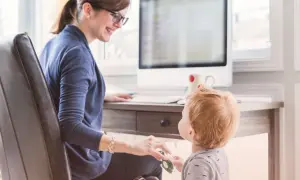En kvinna med glasögon ler mot sitt barn vid ett skrivbord. Hon visar vad betyder flexibel genom att balansera arbete och familjeliv.