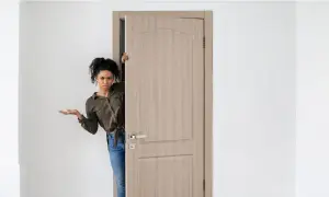 Mujer asomándose por una puerta de madera clara, con expresión de duda. Los colores de puertas varían, pero esta es de un tono neutro.