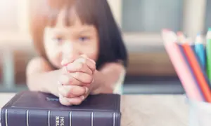 Dziecko z modlącymi się dłońmi na Biblii, obok kolorowe kredki. Nauka 10 przykazań bożych dla dzieci.