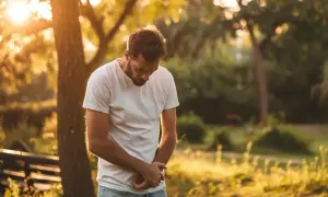 Mężczyzna w białej koszulce trzyma się za brzuch, zgięty w pół. Słońce przebija się przez drzewa, tworząc ciepłą, złotą poświatę. Może to być symbol bólu związanego z chorobą Leśniowskiego-Crohna.
