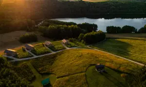 Idealny weekend w Polsce: domki glampingowe nad jeziorem, otoczone zielenią i polami. Słońce zachodzi, rzucając złote światło na krajobraz.