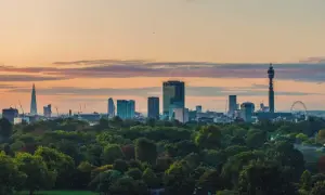 London im Sommer: Die Skyline mit dem Shard und dem BT Tower ragt über einem grünen Park auf, während die Sonne den Himmel in warme Farben taucht.