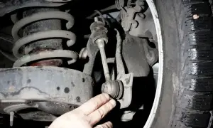 Uszkodzony łącznik stabilizatora objawy: stukanie w zawieszeniu, luzy, nierówna jazda. Widać element zawieszenia i rękę mechanika.