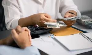 Dłonie liczą banknoty dolarowe, przygotowując dodatek do wynagrodzenia. Obok leży kalkulator i dokumenty.