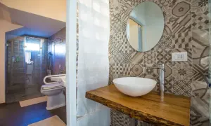 Bagno piccolo con antibagno, doccia in vetro, WC e lavabo su mensola in legno grezzo. Piastrelle geometriche e specchio rotondo.