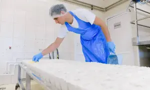 Pracownik w czepku i fartuchu, w rękawiczkach, wykonuje pracę na akord, czyszcząc dużą bryłę sera żółtym narzędziem.