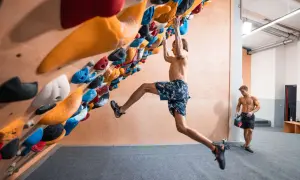 Ein Mann klettert an einer Boulderwand, während ein anderer Mann daneben steht. Der Unterschied zwischen Klettern und Bouldern wird hier deutlich: keine Seile, nur Griffe und eine Matte.