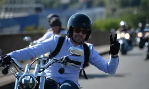 Mężczyzna w kasku i niebieskiej koszuli jedzie na motocyklu i pokazuje znak pokoju.