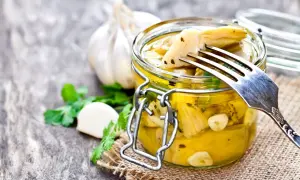 Un barattolo di carciofini sott'olio alla contadina, con aglio e prezzemolo. Una forchetta solleva un carciofino, pronto per essere gustato.