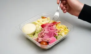 En hand tar en gaffel ur en plastlåda med pastasallad med skinka och fetaost, majs, tomat och dressing.